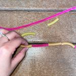A person is putting a piece of tube pasta onto a pink pipe cleaner. Two pieces of pasta have already been threaded and two more are waiting to be added to the chain.