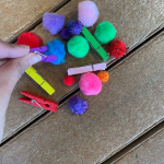 A person is picking up a small, craft pom-pom with a small clothspin.
