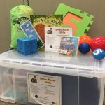School Readiness Kit: a large plastic box with a contents label titled "Gross Motor Skills". On top of the box are a 12 ft. parachute green bag, a blue block that has activities such as touch toes and skip, a brochure for Early Learning Kits, a book titled "You Are a Lion!". a green foam square with an orange number 5, a CD called "Kids in Action, and four balls of various sizes (orange, red, dark blue and light blue)